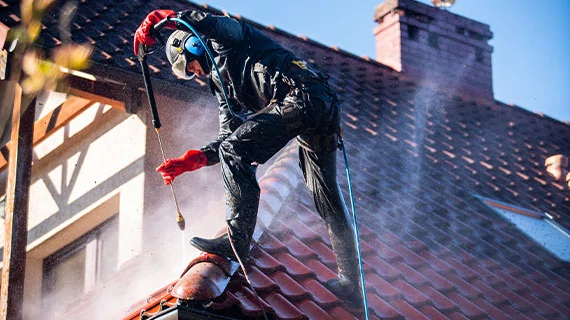 Ein Facharbeiter reinigt ein Hausdach mit einem Hochdruckreiniger. Auf den Dachziegeln ist deutlich der starke Wasserstrahl zu sehen, der Schmutz und Ablagerungen entfernt.